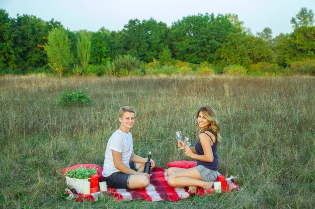 Hermosa joven pareja amorosa feliz en picnic acostado en cuadros en el campo en un día soleado de verano disfrutando, sosteniendo vino y descansando. mirando a la cámara y sonriendo.