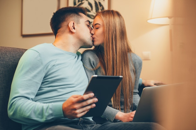 Hermosa joven pareja besándose mientras se apoya en un sofá sosteniendo una computadora portátil y una mesa en su casa.