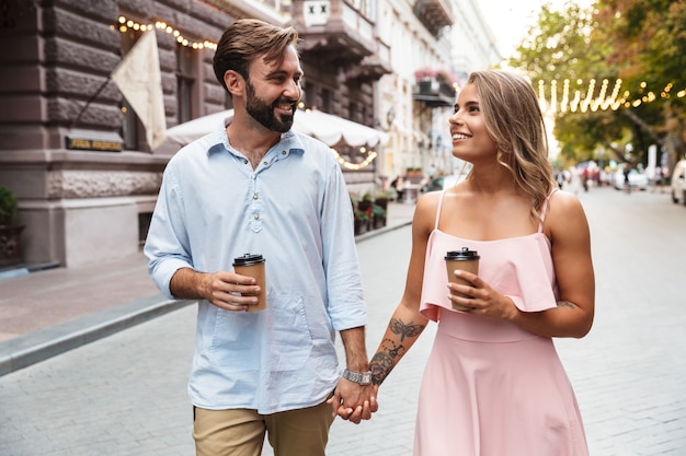 Hermosa joven pareja caminando al aire libre por la calle de la ciudad, tomados de la mano, bebiendo café para llevar