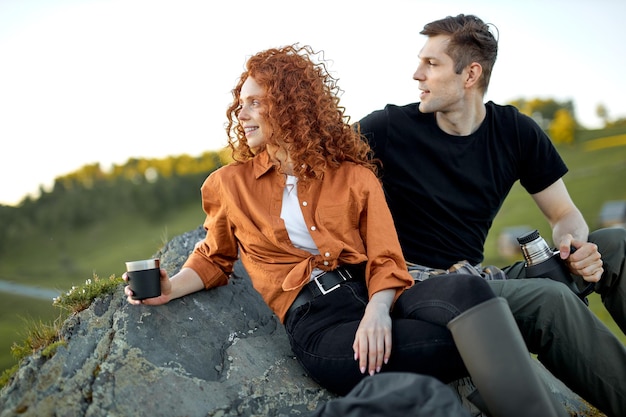 Hermosa joven pareja caucásica sentarse tomando un descanso descansando en las montañas bebiendo té del termo ...