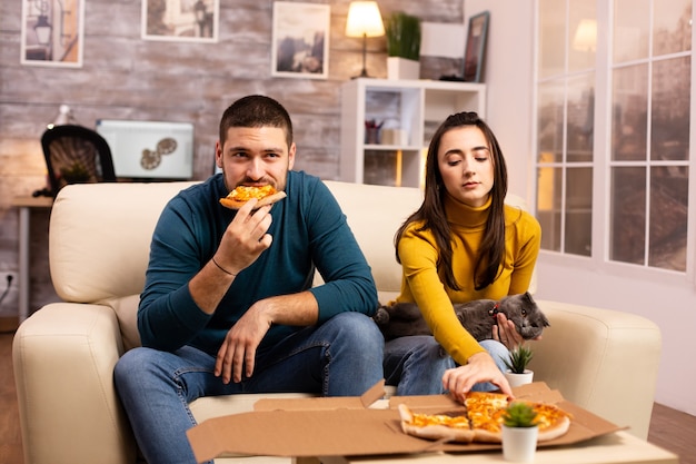 Hermosa joven pareja comiendo pizza mientras ve la televisión en la sala de estar sentado en el sofá