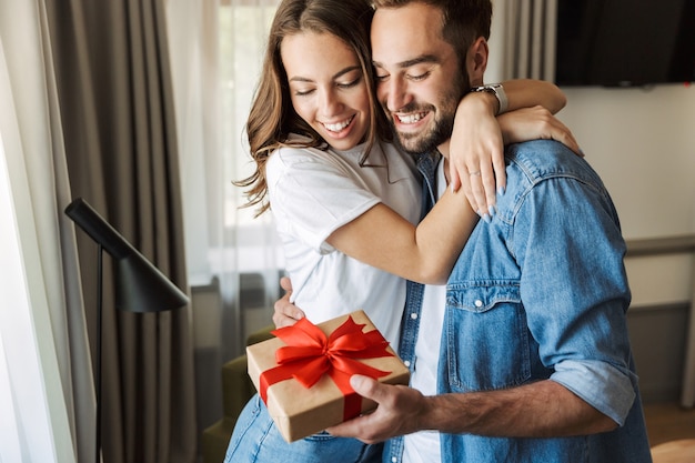 Hermosa joven pareja enamorada en casa, celebrando con un intercambio de caja de regalo, abrazos