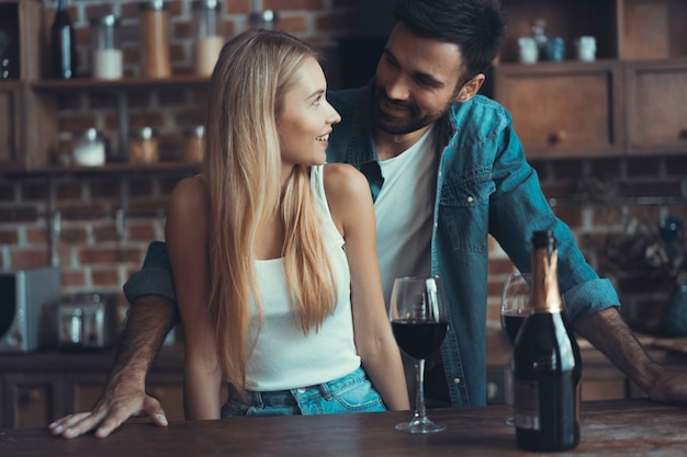 Hermosa joven pareja está bebiendo vino y sonriendo mientras cocina en la cocina de su casa.