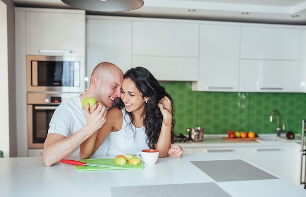 Hermosa joven pareja está hablando, mirando a cámara y sonriendo mientras se cocina en la cocina.