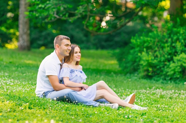 Hermosa joven pareja está pasando un buen rato sentados en el césped del parque abrazándose mirando a lo lejos