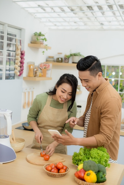 Hermosa joven pareja está usando un teléfono celular y sonriendo mientras cocina en la cocina de casa