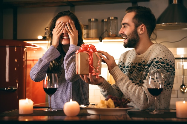 Hermosa joven pareja feliz pasar una velada romántica juntos en casa, bebiendo vino tinto, hombre dando un regalo