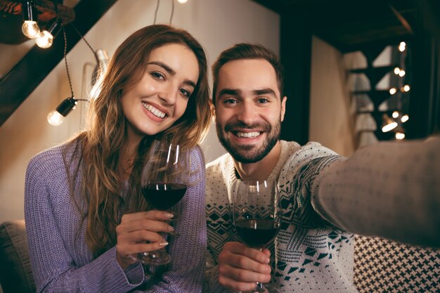 Hermosa joven pareja feliz pasar una velada romántica juntos en casa, bebiendo vino tinto, tomando un selfie