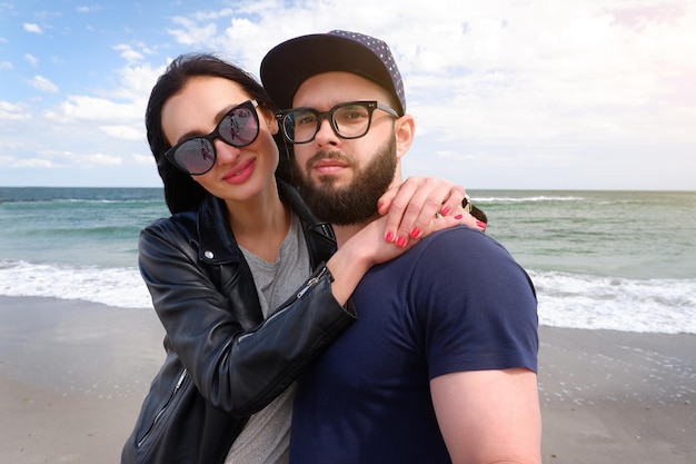 Hermosa joven pareja de hipsters en ropa de moda en el fondo del mar. Chico barbudo camiseta azul gorra de béisbol con gafas de sol sonriendo felices amantes en la playa junto al mar.