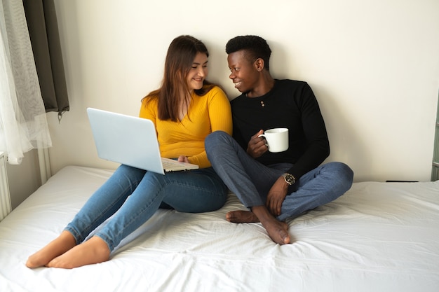 Hermosa joven pareja hombre africano y mujer caucásica sentarse en la cama con un portátil