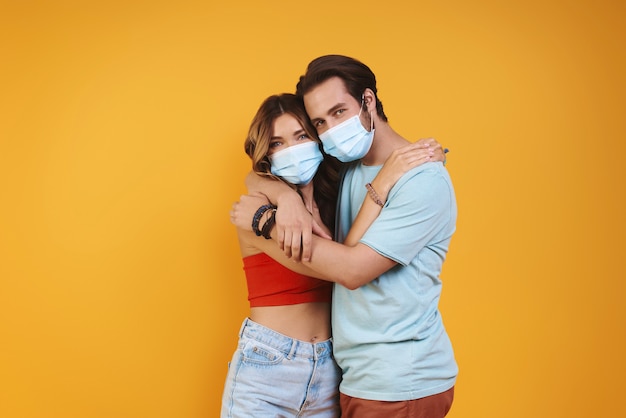Foto hermosa joven pareja en máscaras protectoras abrazando mientras está de pie contra el fondo amarillo