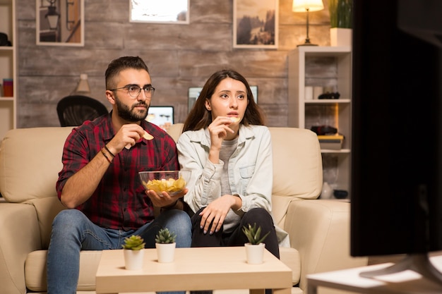 Hermosa joven pareja mirando preocupado en la televisión mientras ve una película. Pareja comiendo patatas fritas sentado en el sofá.