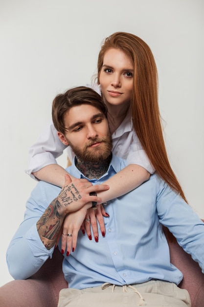 Hermosa joven pareja de moda Mujer elegante pelirroja de moda bonita abraza a un apuesto hombre hipster con tatuajes en un estudio blanco