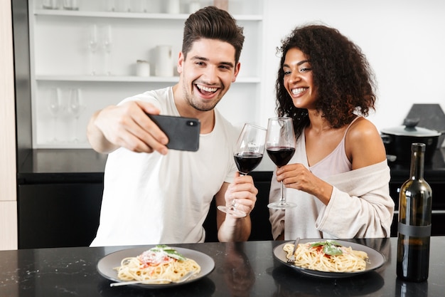 Hermosa joven pareja multiétnica con una cena romántica en casa, bebiendo vino tinto y comiendo pasta, brindando, tomando un selfie