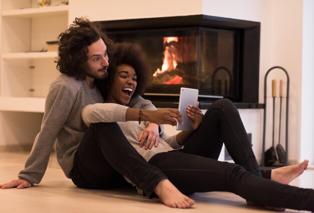 hermosa joven pareja multiétnica usando una tableta en el piso de su casa de lujo frente a la chimenea en el día de otoño