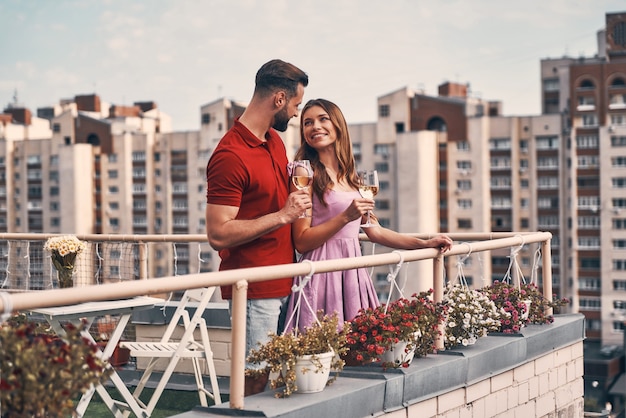 Hermosa joven pareja en ropa casual abrazando y mirando a otro lado con una sonrisa mientras está de pie en el patio de la azotea al aire libre