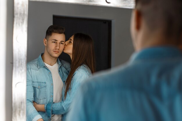 Hermosa joven pareja en ropa elegante de mezclilla abrazándose cerca de un espejo vintage con luces