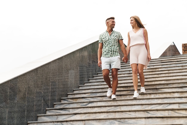 Hermosa joven pareja en ropa de verano sonriendo y tomados de la mano juntos mientras camina por las escaleras de la ciudad