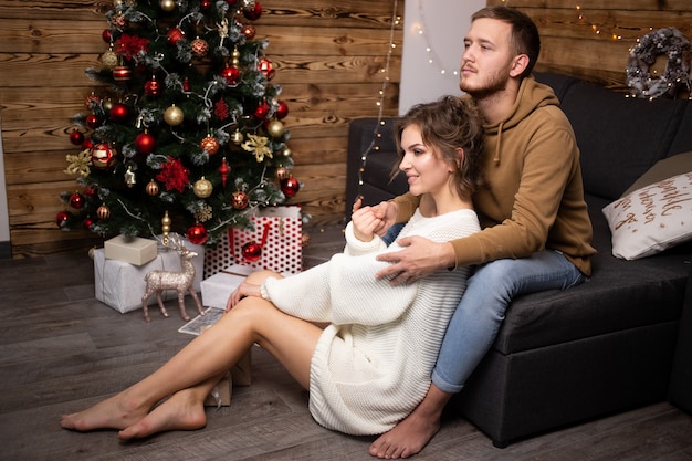 Hermosa joven pareja sentada en el piso de la sala junto a un árbol de Navidad muy bien decorado