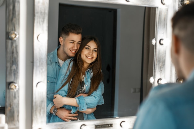 Hermosa joven pareja con sonrisa en ropa de jeans abrazándose cerca de un espejo vintage con luces