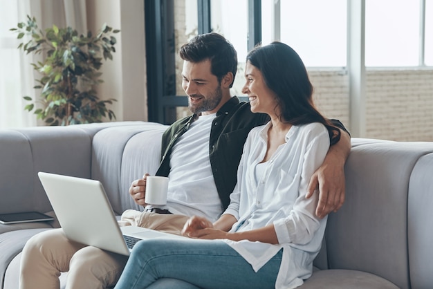 Hermosa joven pareja tomando café y mirando la computadora portátil mientras está sentado en el sofá en casa