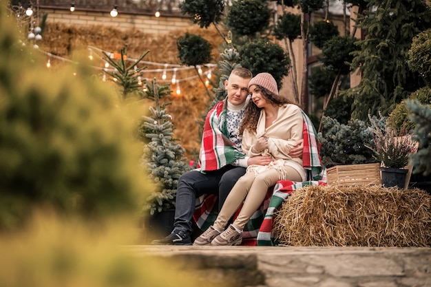 Una hermosa joven pareja vestida de blanco está sentada cerca de un árbol de Navidad en el jardín. Hombre y mujer felices, romance, celebración de Navidad, diversión, amor.