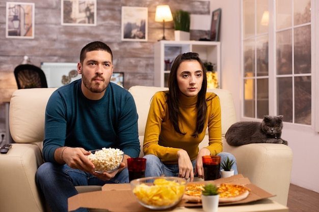 Hermosa joven pareja viendo la televisión y comiendo comida rápida para llevar en la sala de estar