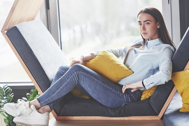 Hermosa joven pasa su tiempo en el área de recreación. Se sienta y se divierte en el sillón en forma de hexágono con almohadas cerca de la ventana.