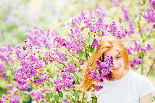 Hermosa joven pelirroja adolescente de pie en arbustos de lilas en flor y mirando a la cámara