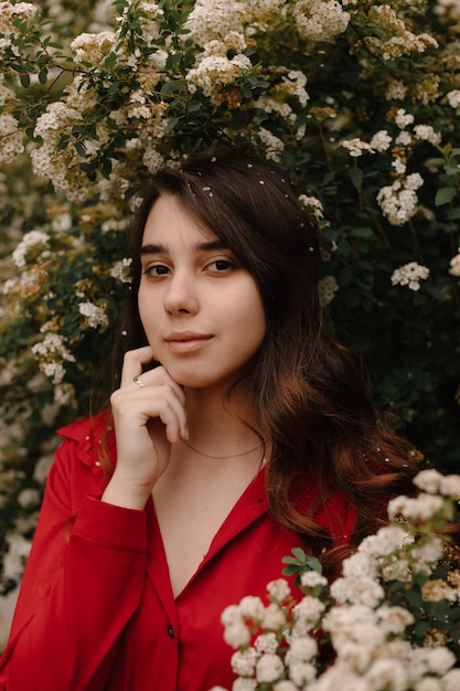 Hermosa joven pelirroja en una blusa roja en la primavera cerca de arbustos en flor