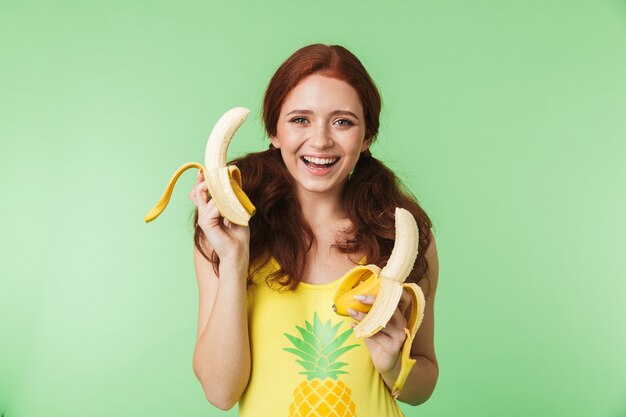 Hermosa joven pelirroja emocionada posando aislada sobre fondo de pared verde con frutas de plátano.