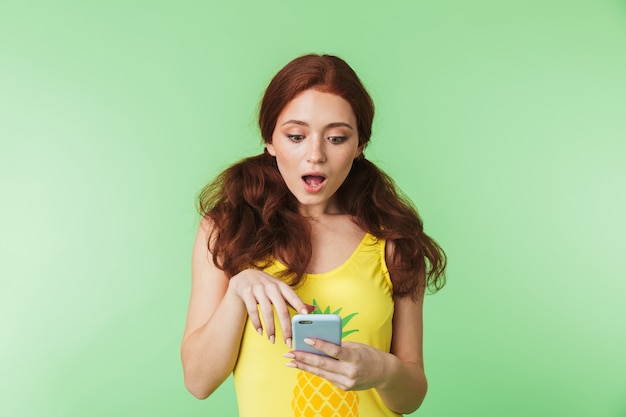 Hermosa joven pelirroja emocional conmocionada posando aislada sobre fondo de pared verde mediante teléfono móvil.