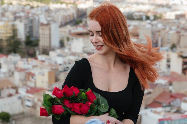 Hermosa joven pelirroja enamorada, mira tiernamente las rosas rojas en sus brazos. Día de San Valentín