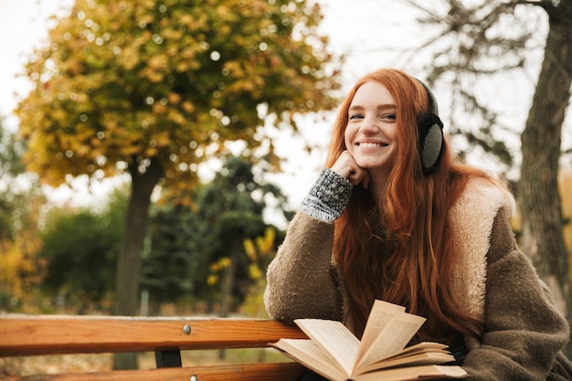 Hermosa joven pelirroja escuchando música con headpones mientras está sentado en un banco, leyendo un libro