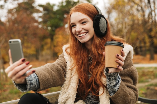 Hermosa joven pelirroja escuchando música con headpones mientras está sentado en un banco, tomando un selfie