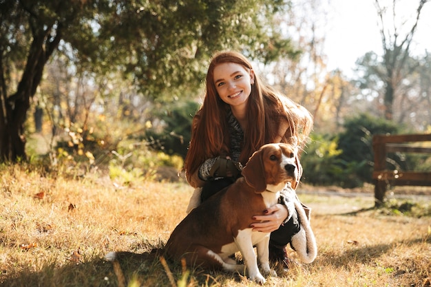 Foto hermosa joven pelirroja escuchando música con tocados mientras juega con su perro en el parque