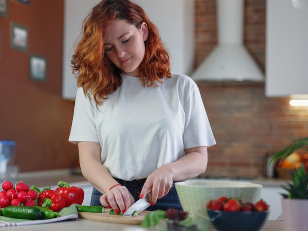 Hermosa joven pelirroja está preparando ensalada de verduras en la cocina