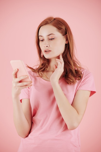 Hermosa joven pelirroja leyendo mensajes de texto en el teléfono inteligente, aislado en rosa