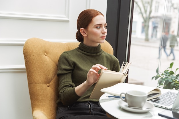 Hermosa joven pelirroja relajante en la mesa de café en el interior, tomando café, leyendo un libro