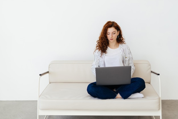 Hermosa joven pelirroja sentada en un sofá en casa escribiendo en una laptop