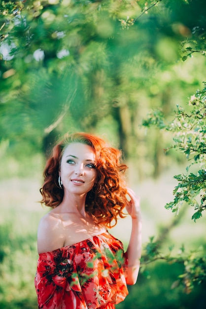 Hermosa joven pelirroja en vestido rojo entre el verde follaje del árbol sonriendo.