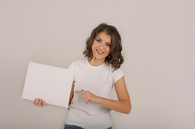 Una hermosa joven de pelo castaño con una camiseta blanca sostiene una hoja de papel blanco en sus manos