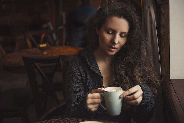 Foto hermosa joven de pelo castaño sentado en un café y bebiendo un capuchino.