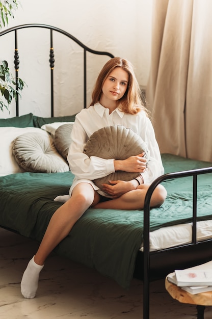 Hermosa joven con el pelo largo con una camisa blanca se sienta en una cama en el dormitorio por la mañana