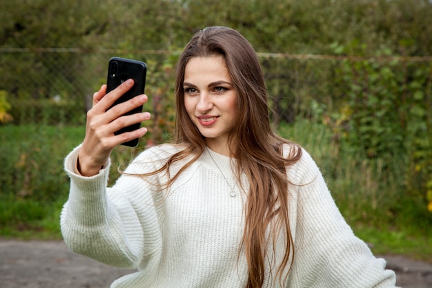 Una hermosa joven de pelo largo con un suéter blanco hace un selfie en el teléfono.