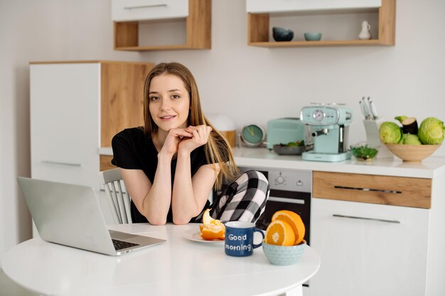 Hermosa joven en pijama trabajando con una laptop en la cocina por la mañana