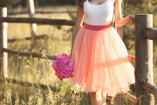 Hermosa joven con piones en la naturaleza al aire libre bajo el sol. Linda mujer afuera con flores en la falda rosa