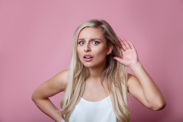 Hermosa joven pone una mano en el oído para escuchar mejor en una pared rosa