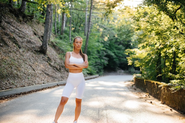 Una hermosa joven posa antes de ejecutar el entrenamiento, en la carretera en un denso bosque, durante la puesta de sol. Un estilo de vida saludable y correr al aire libre.