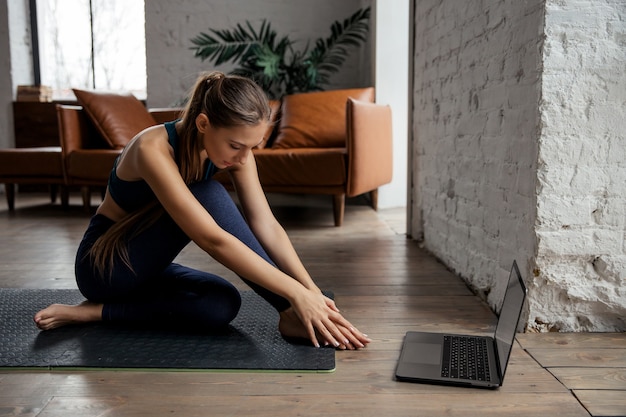 Hermosa joven practicando yoga en casa con ordenador portátil. Concepto de entrenamiento en línea. Foto de alta calidad